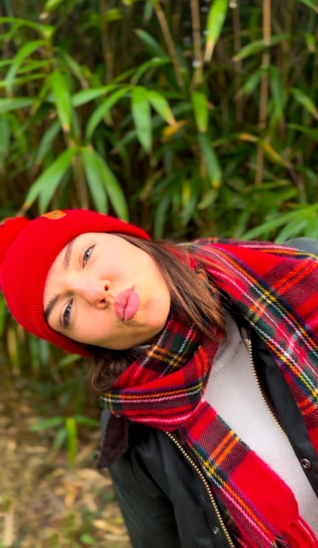 Person wearing a red beanie and plaid scarf in front of green foliage