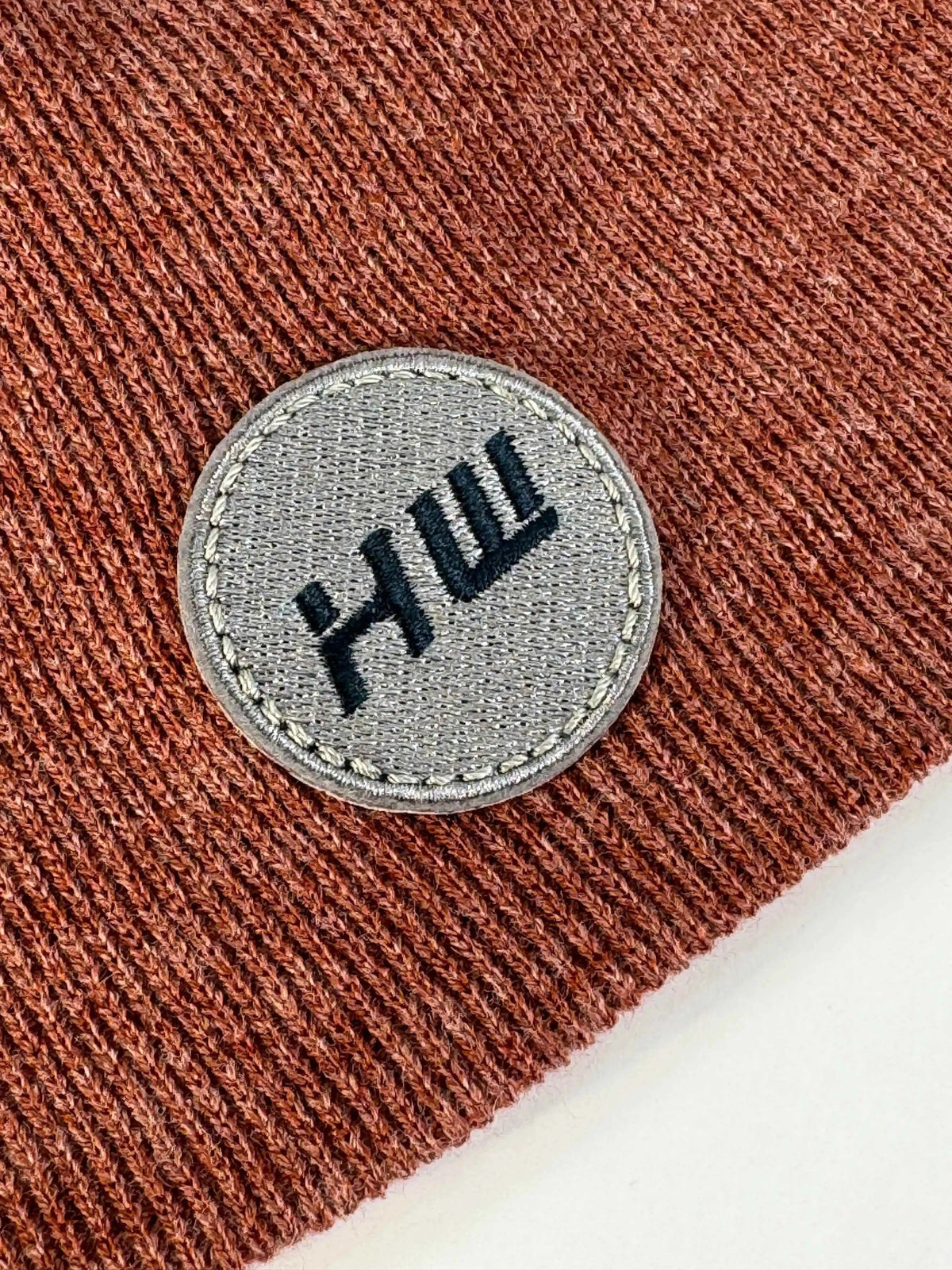Rust-brown knit beanie with stitched round logo patch on front cuff