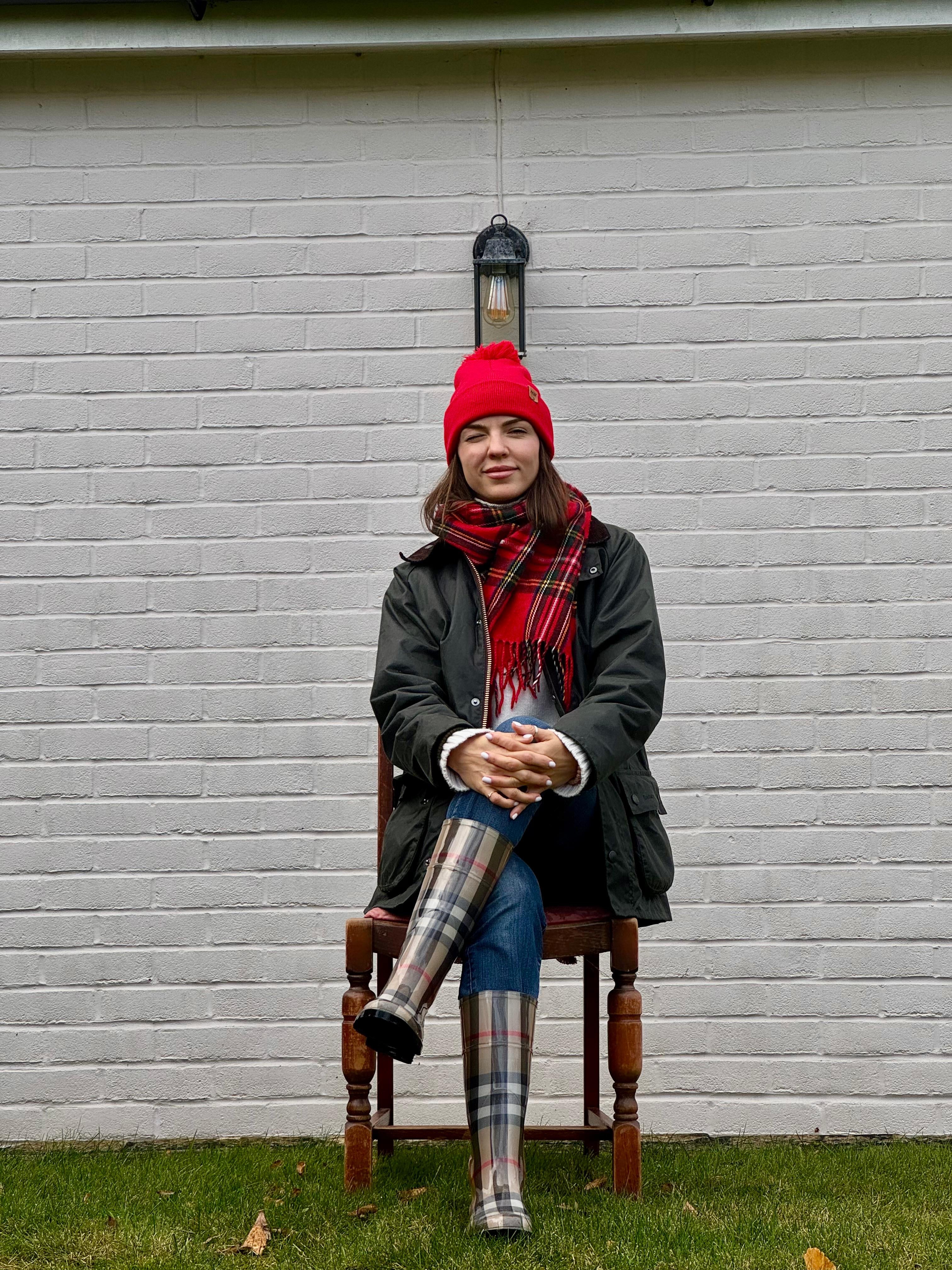 Person wearing a red beanie, dark coat, plaid scarf, and tartan pants sitting on a wooden chair against a gray brick wall.