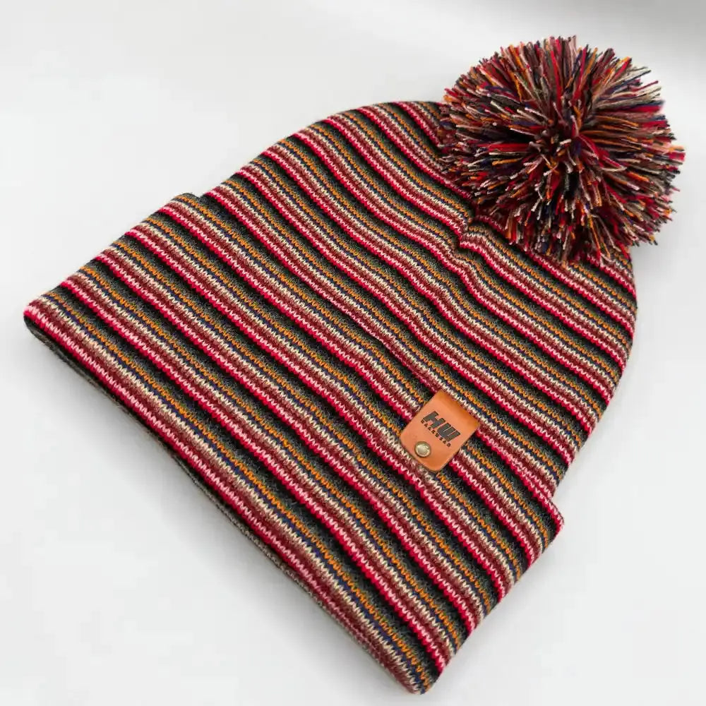 Multicolor striped knit beanie with pom pom and tan logo patch, folded view