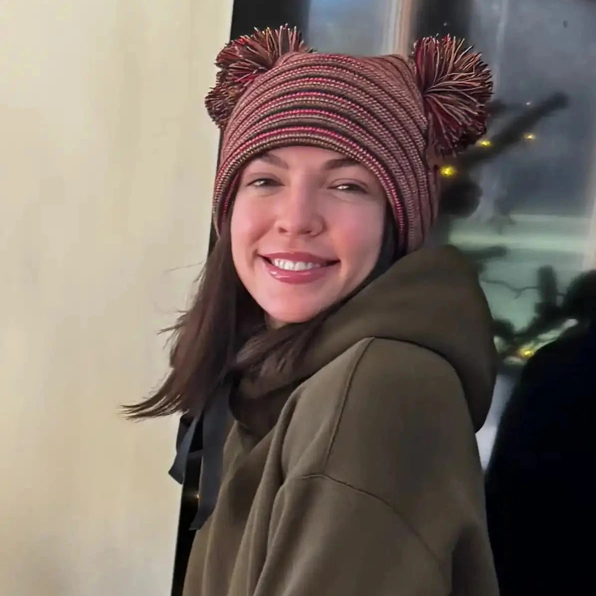 Maroon ribbed beanie with two pom poms and multicolor stripes, worn by a smiling person.