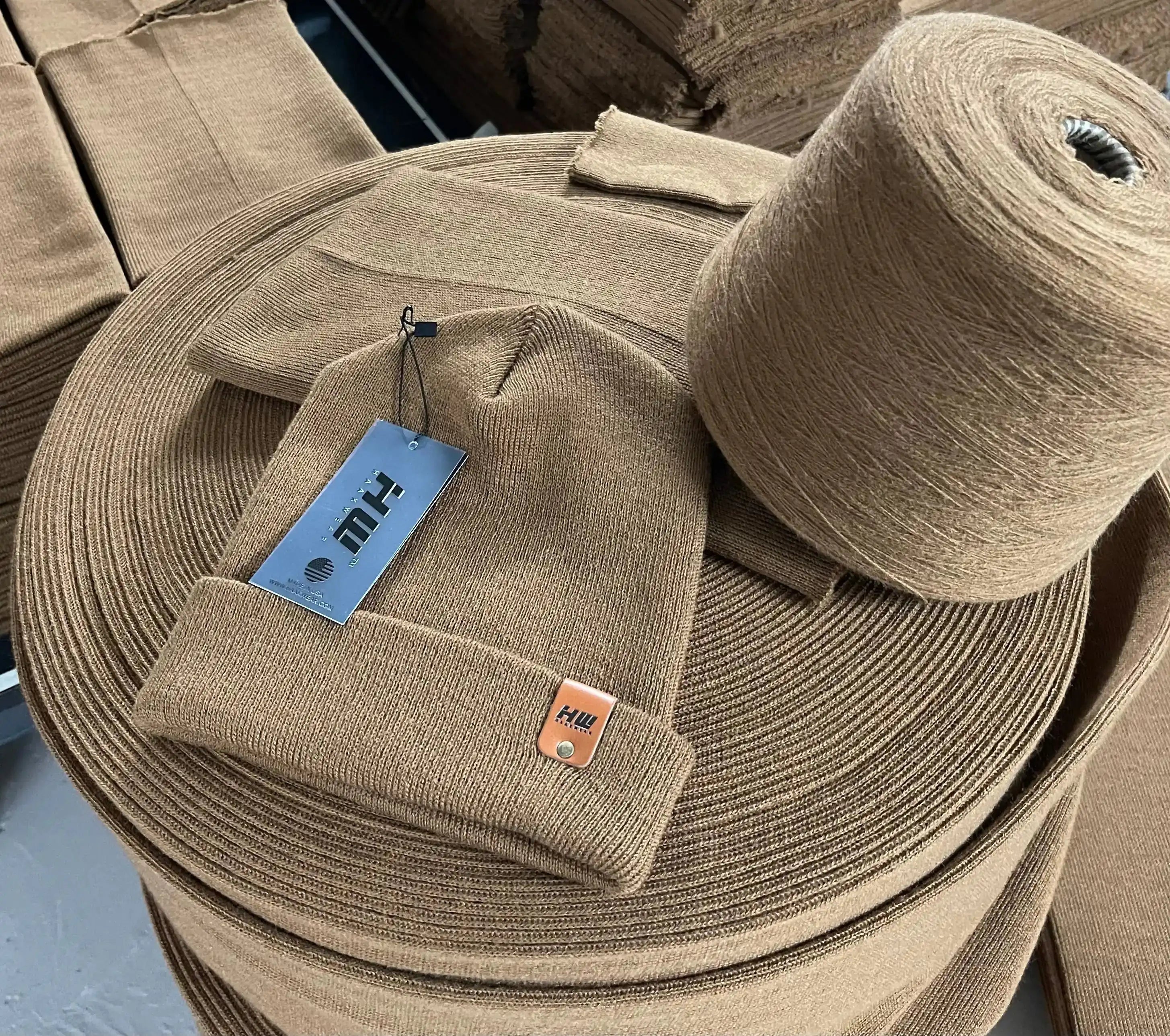 Brown fabric roll with a knit cap and tag, on a background of more fabric rolls showing how beanies are made. 