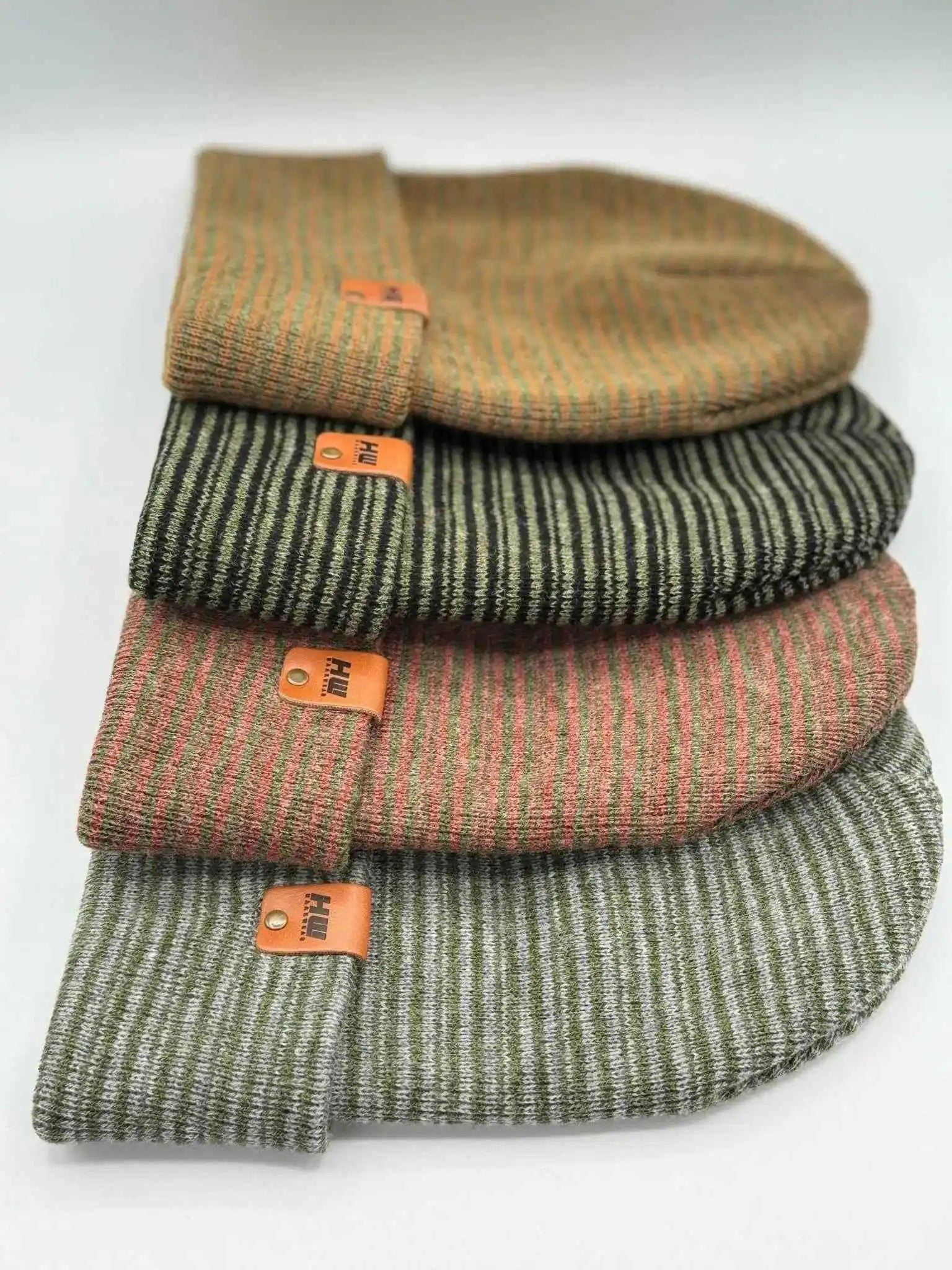 Stack of knit beanies with folded cuffs and orange logo patches, gray-green tones
