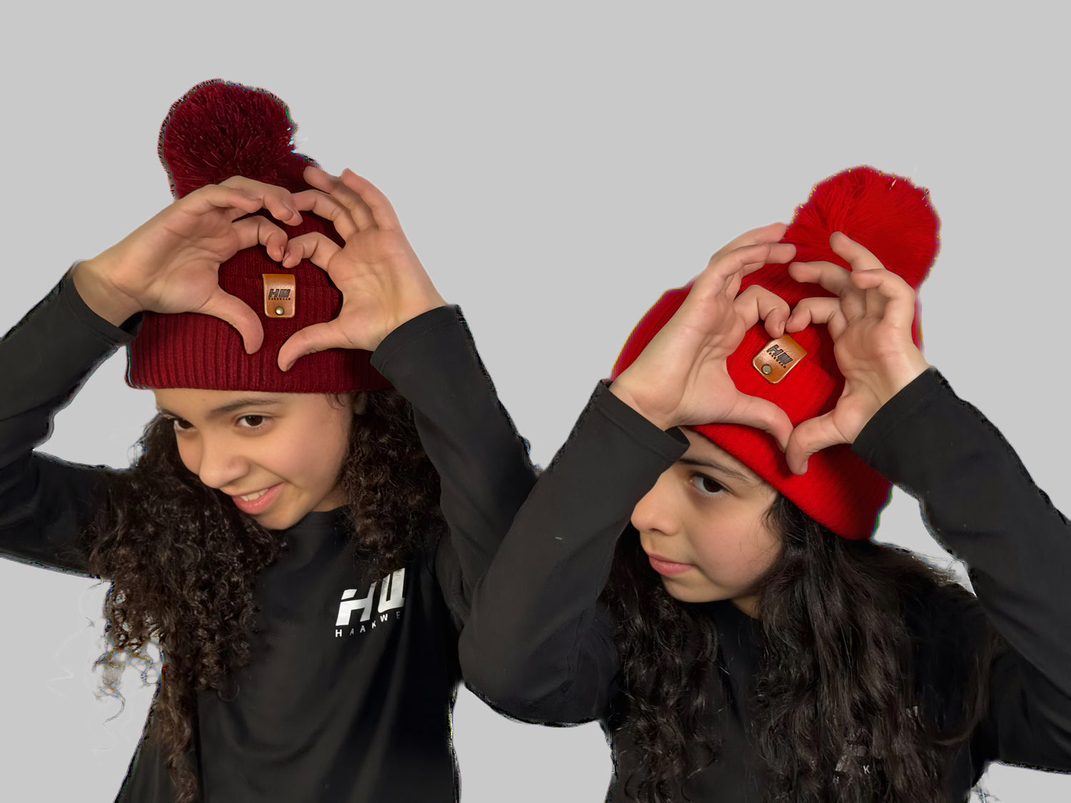 Two children wearing red beanies with pom-poms, making heart shapes with their hands against a gray background. Made in USA beanies with Free shipping 