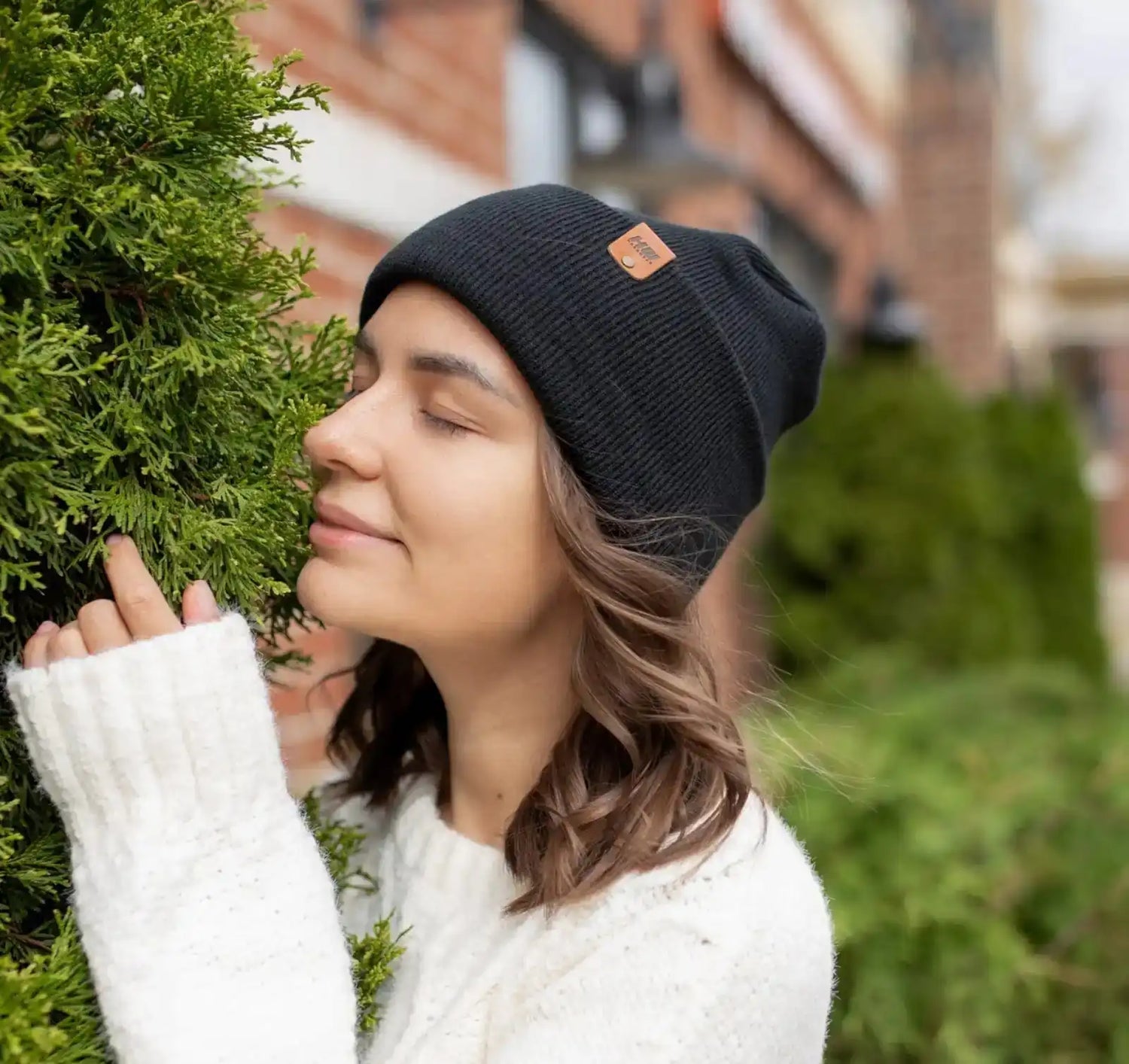 Woman wearing a black beanie with a brand logo, standing next to a green bush.
