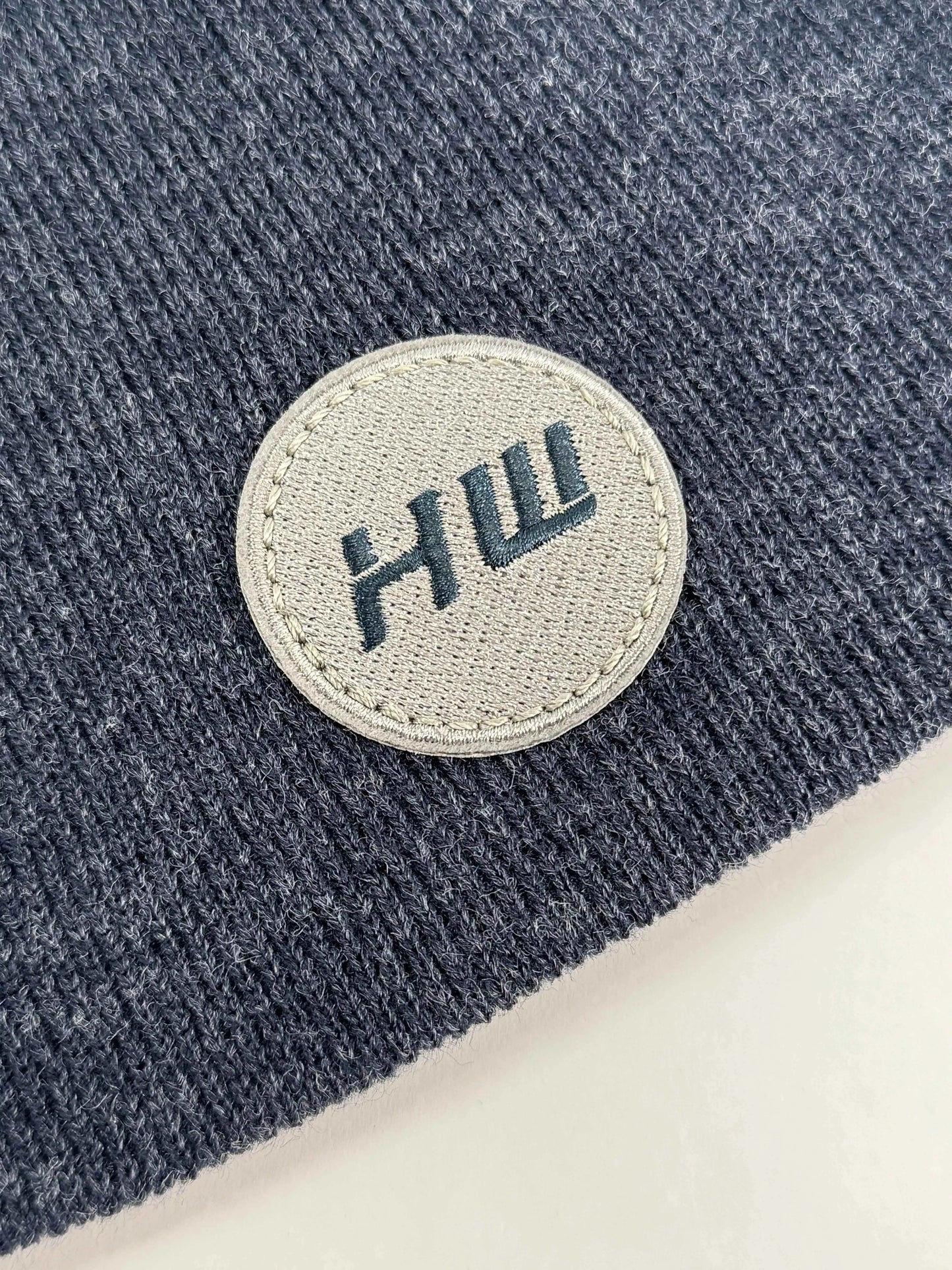 Close-up of denim blue knit beanie with round HUI logo patch.