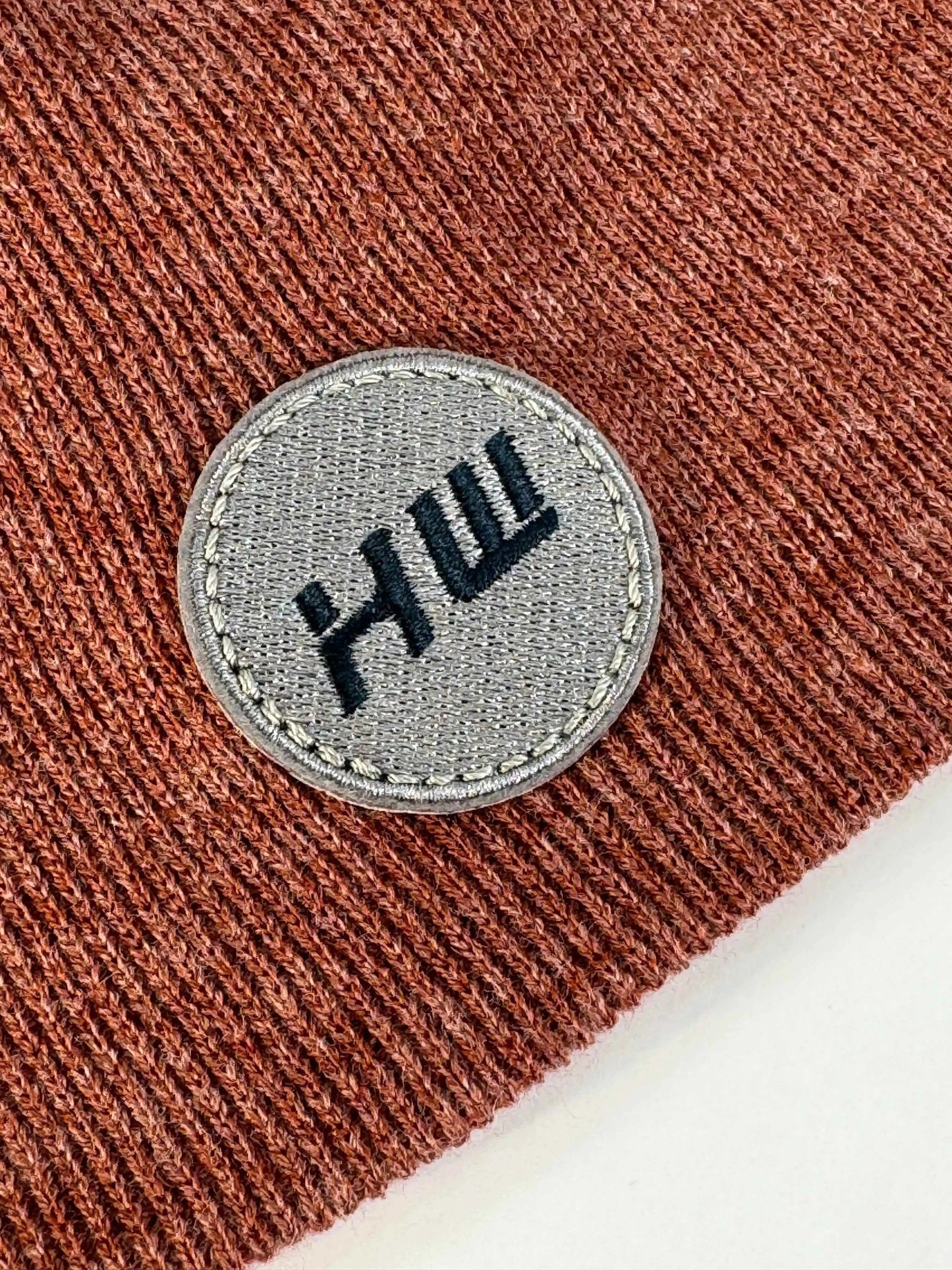 Rust-brown knit beanie with stitched round logo patch on front cuff