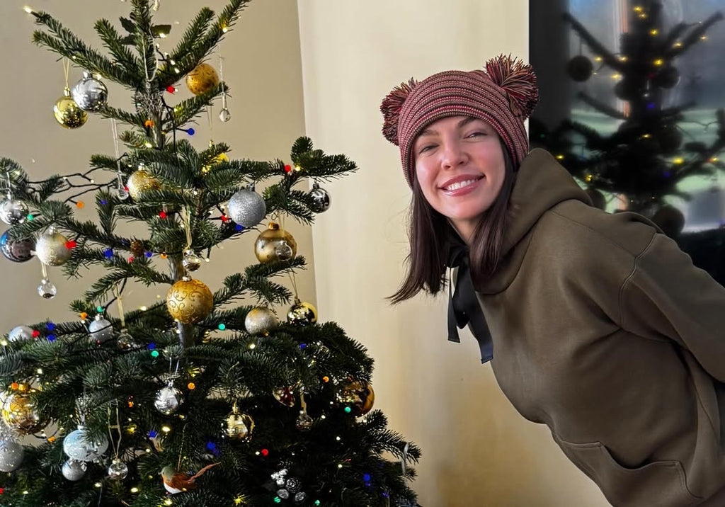 Person wearing a double pom pom beanie made in USA by Haakwear and smiling next to a decorated Christmas tree.