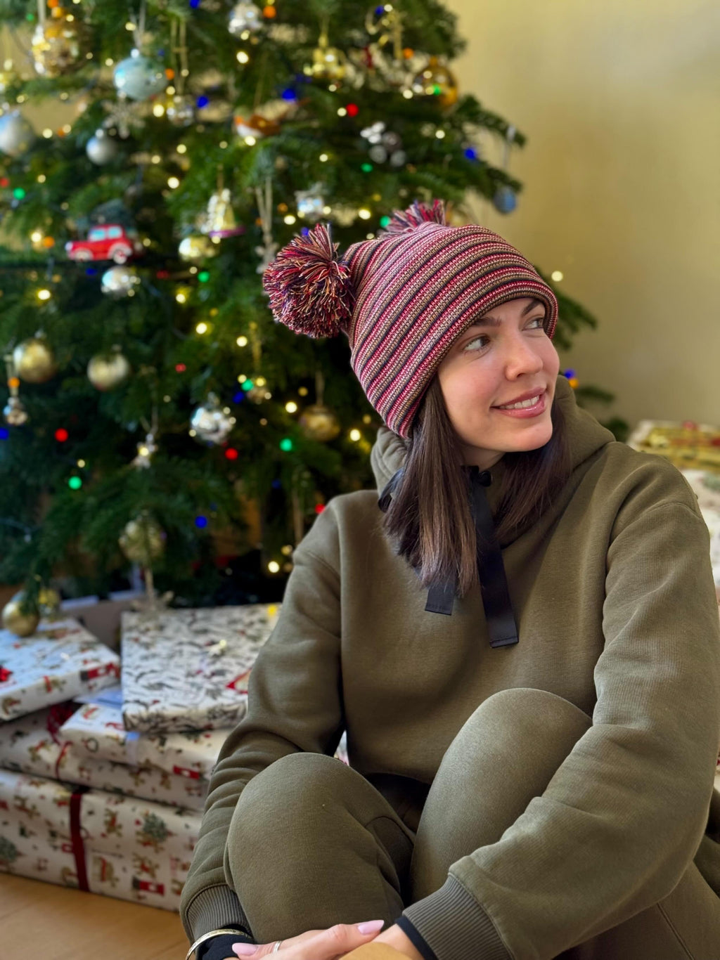 Person wearing a striped multi color beanie made in use with hand made pom-pom in front of a decorated Christmas tree.