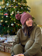 Person wearing a striped multi color beanie made in use with hand made pom-pom in front of a decorated Christmas tree.