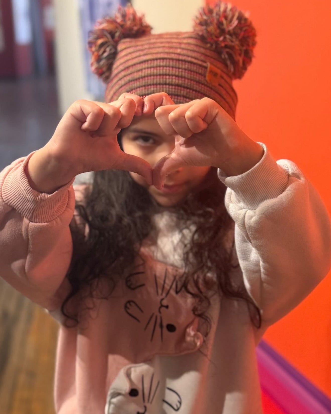 Child wearing a pink sweater with a cat design and a striped hat with pom-poms, making a heart shape with their hands. the beanie is made in USA by haakwear for the haakwear kids beanie collection 