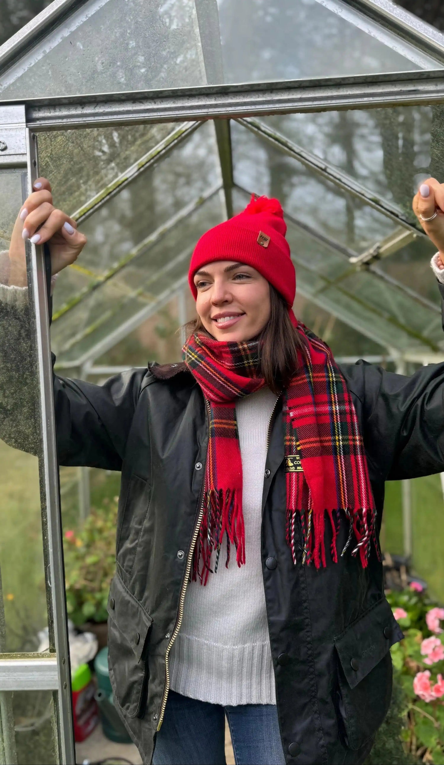 women wearing a red beanie and plaid scarf standing inside a greenhouse. 